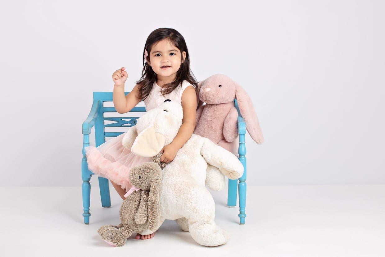 2 year old girl on blue bench with two stuffed bunnies posing for photoshoot