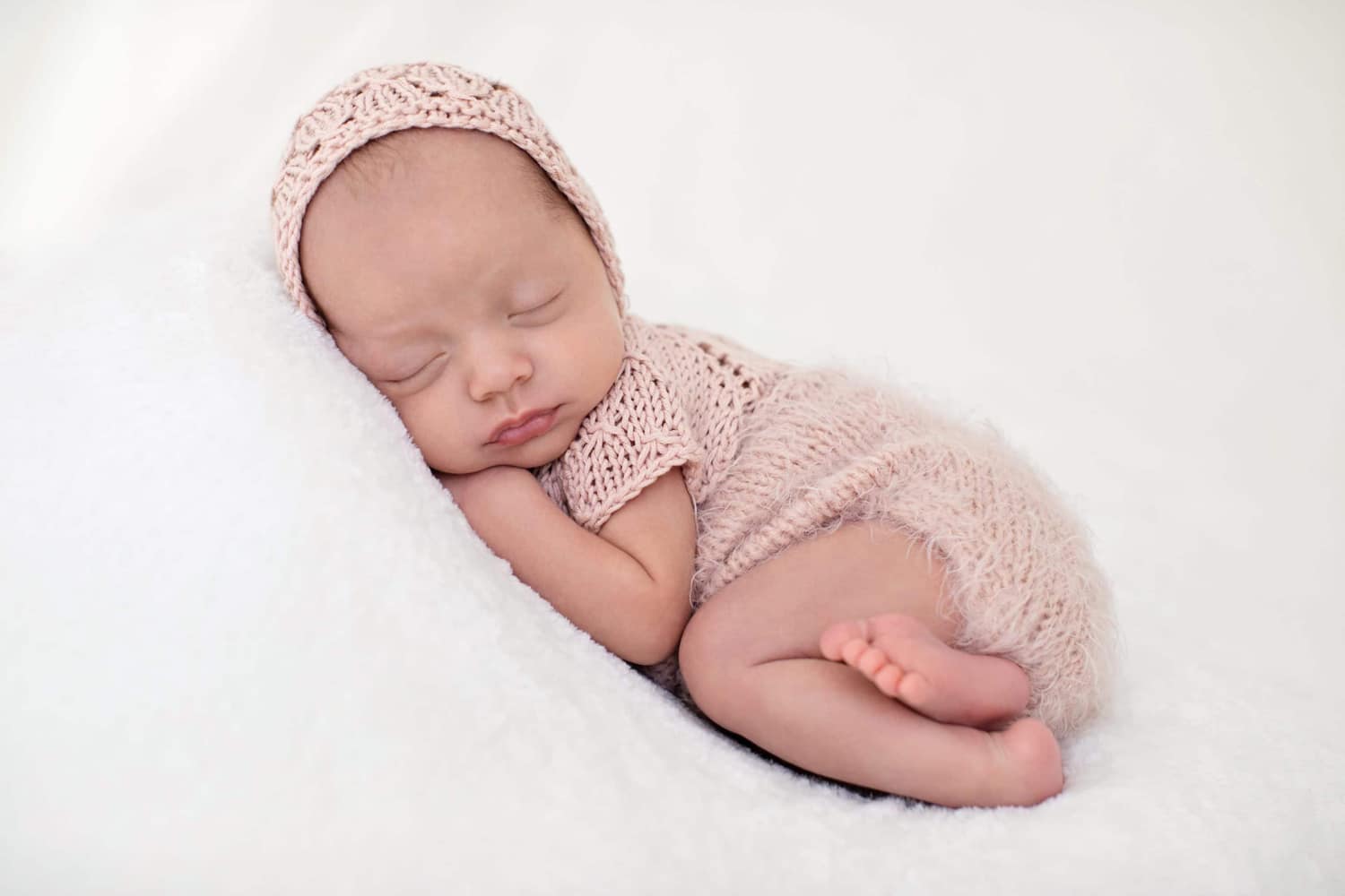 A baby snuggles on a white pillow wearing a pink knit outfit.