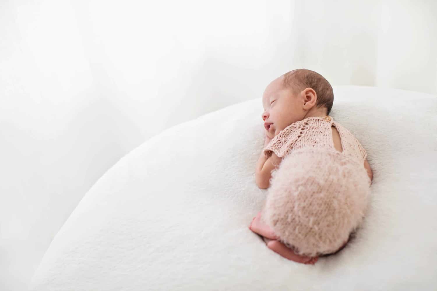 A baby snuggles on a white pillow wearing a pink knit outfit.