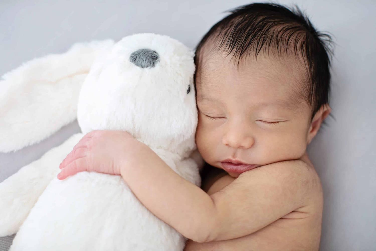 A newborn baby hugs a white stuffed rabbit.