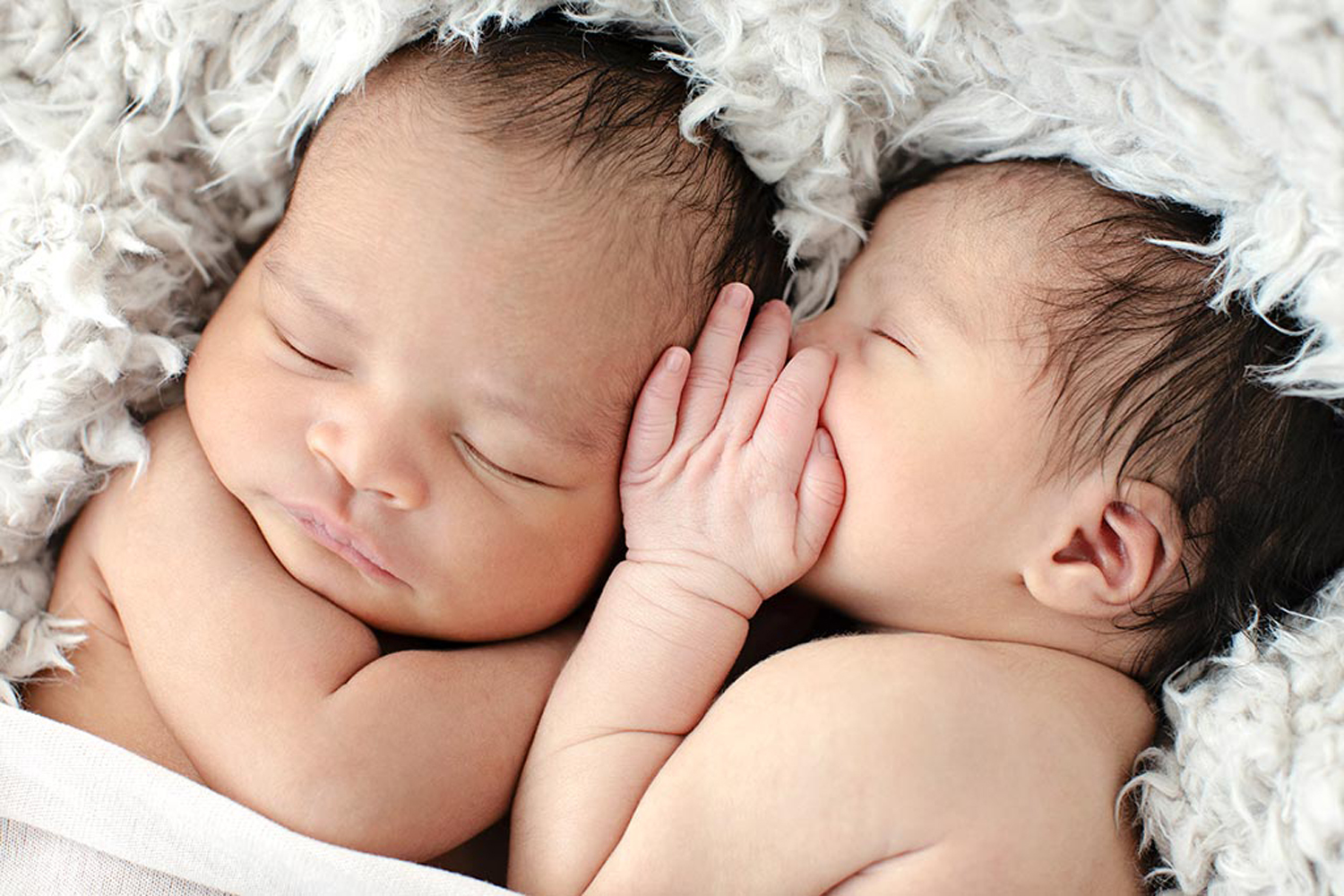 Two newborn babies cuddle on a fluffy white material.