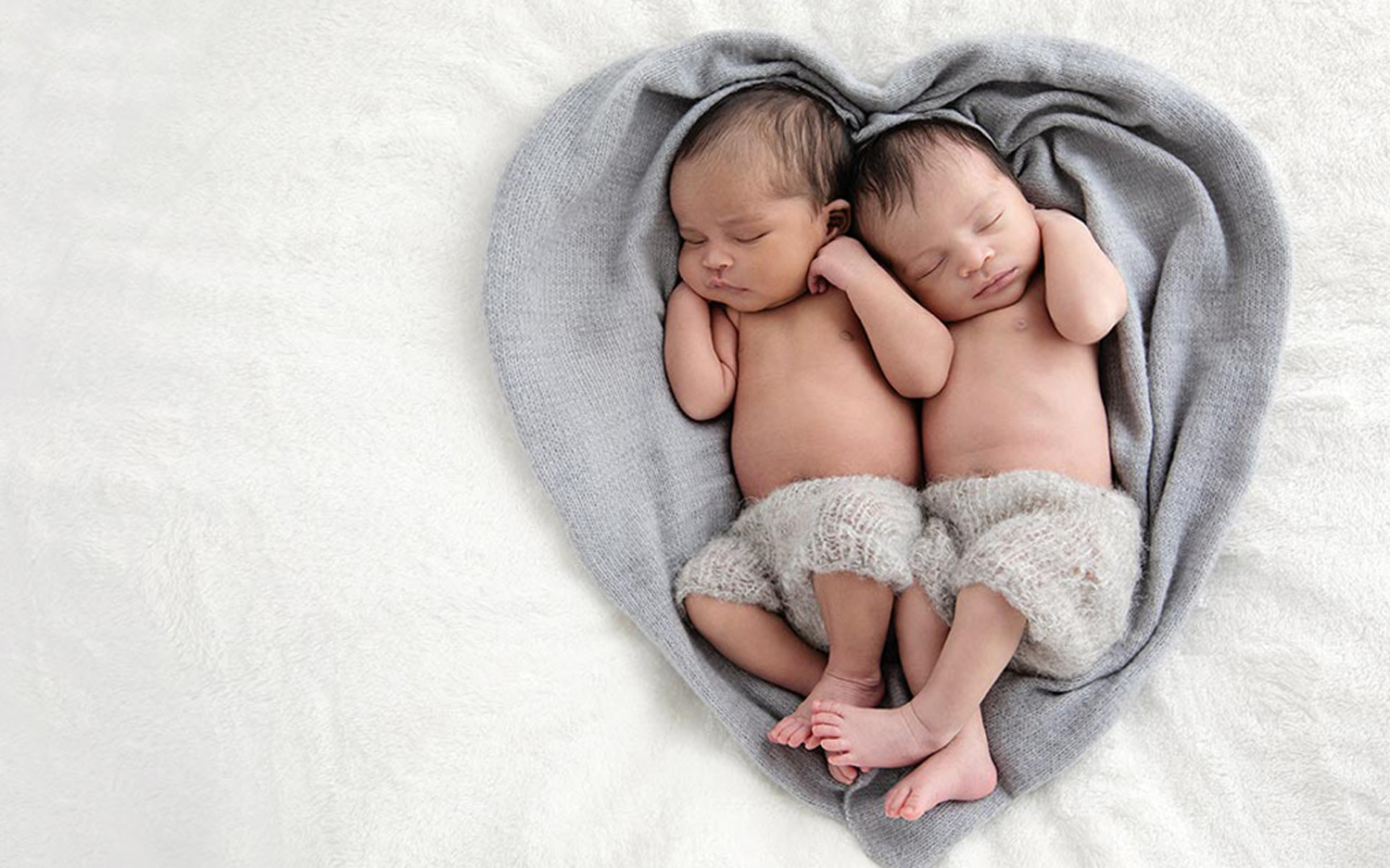 Two newborn babies cuddle on a heart-shaped gray fabric.