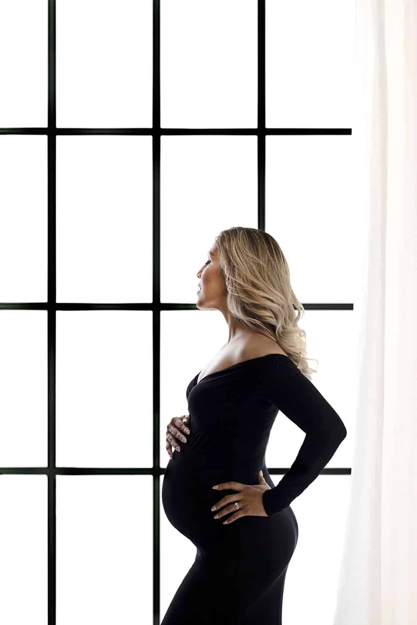 A pregnant woman in a black dress in front of a window.