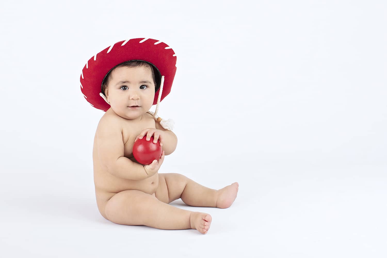 A six month old wearing a red cowboy hat holds a red ball.