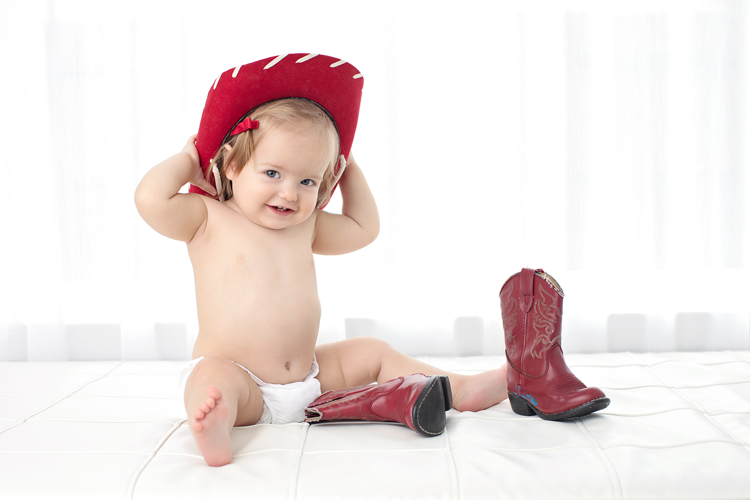 A charming baby sits in a red cowboy hat with boots